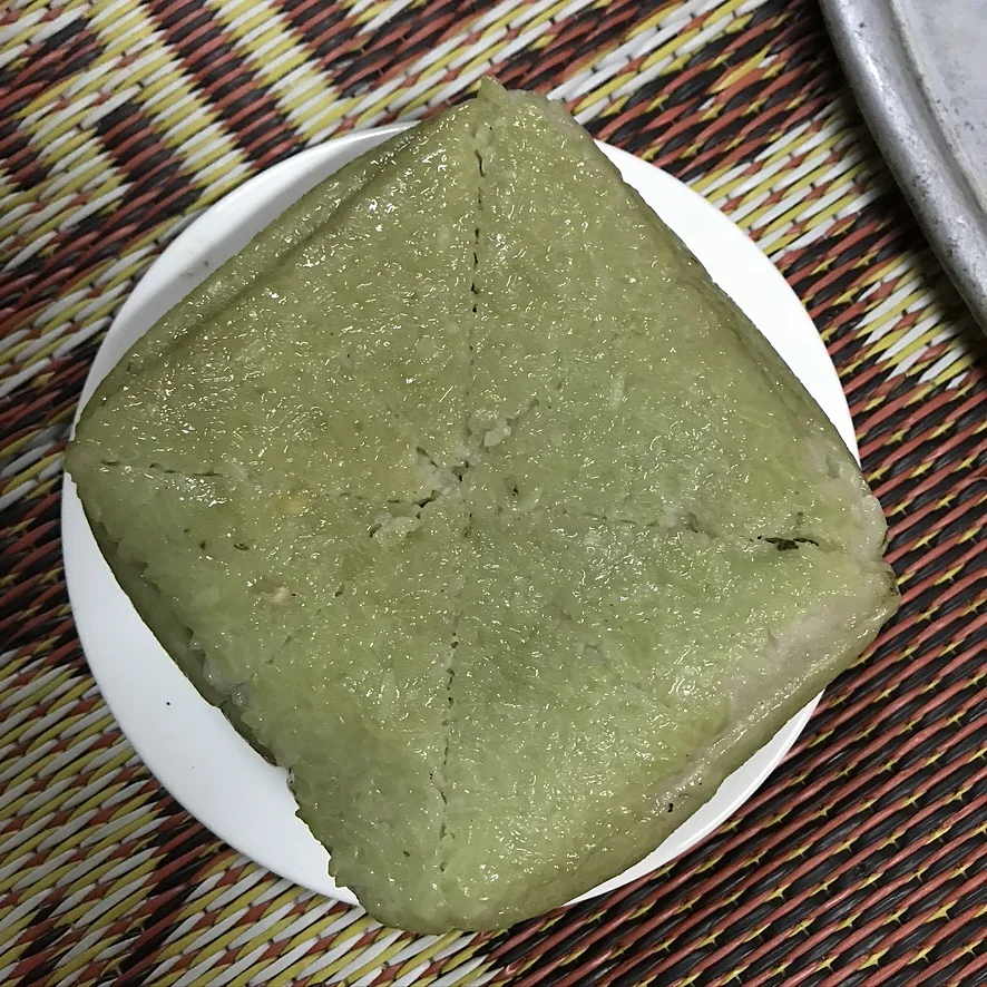 Chiếc bánh chưng vuông vức đã bóc lá, vỏ xanh đều, đặt trên đĩa trắng trên chiếu hoa
