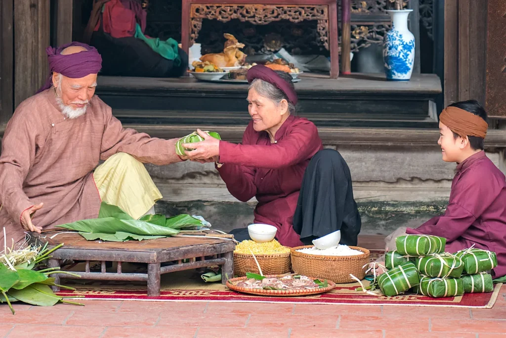 Ông bà và cháu mặc áo truyền thống ngồi gói bánh chưng trước hiên nhà, phía sau là bàn thờ bày lễ ngày Tết