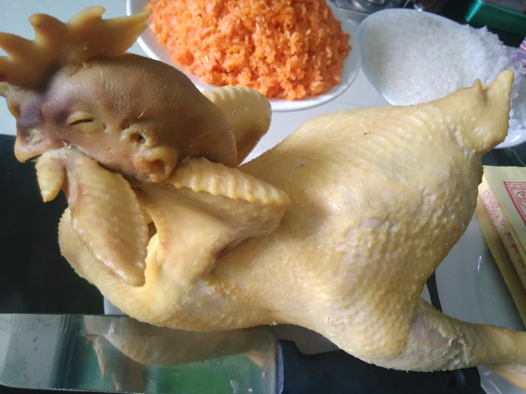 Gà trống luộc còn nguyên mào, cánh gập sát thân, đặt cạnh đĩa xôi gấc trên bàn cúng