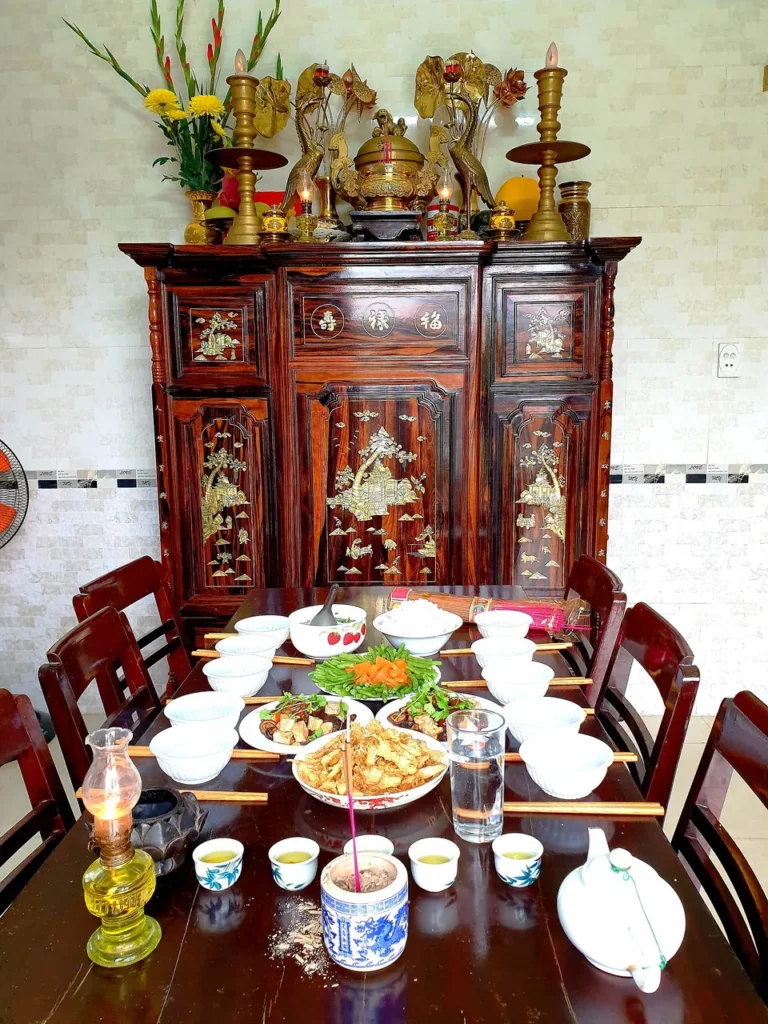 Bàn cỗ cúng Tết bày nhiều bát đĩa thức ăn, chén nước và đèn dầu trước tủ thờ gỗ khảm