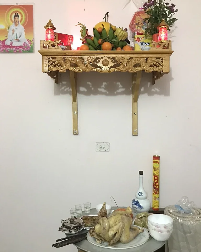 Bàn thờ treo trên tường bày mâm ngũ quả chuối xanh, bưởi, phật thủ, cam cùng hoa cúc và đèn thờ; phía dưới là mâm cúng có gà luộc, giò, xôi, chén rượu và bình rượu