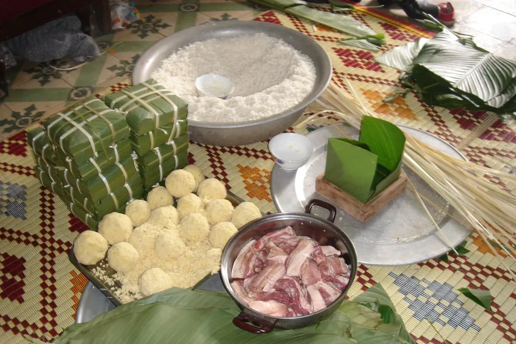 Gạo nếp, đậu xanh nắm, thịt ba chỉ, lạt, lá dong và khuôn gỗ trên chiếu, cạnh chồng bánh chưng đã gói