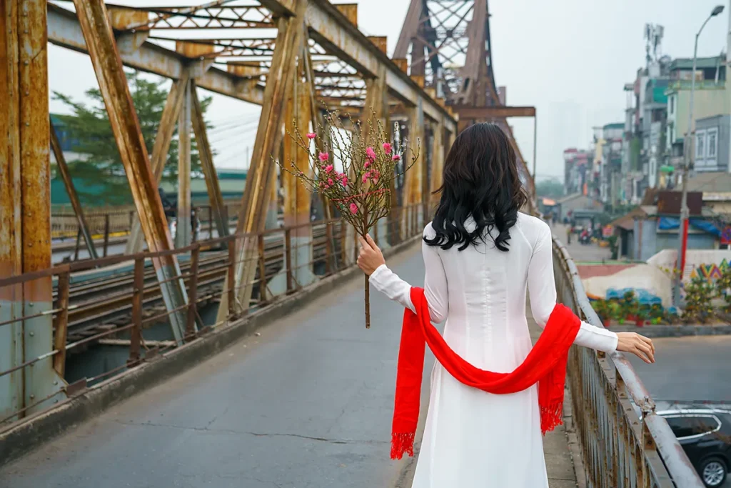 Cô gái mặc áo dài trắng, khoác khăn đỏ, cầm cành đào đi trên cầu Long Biên ngày Tết